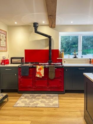 Shaker kitchen featuring a glossy red range with a black stovepipe, dark cabinets, a window overlooking trees, and a patterned rug on a wooden floor.
