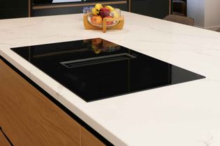 Modern kitchen with a sleek black induction cooktop on a spacious white marble island. A wooden fruit basket adds a touch of colour and warmth.