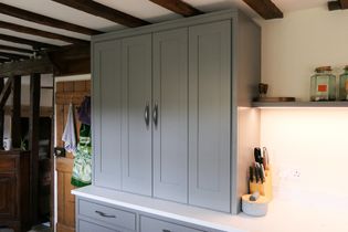 Shaker kitchen with grey countertop cabinet, exposed wooden beams, and a rustic door. To the right of it is a knife set, two jars, and soft lighting for a cozy feel.