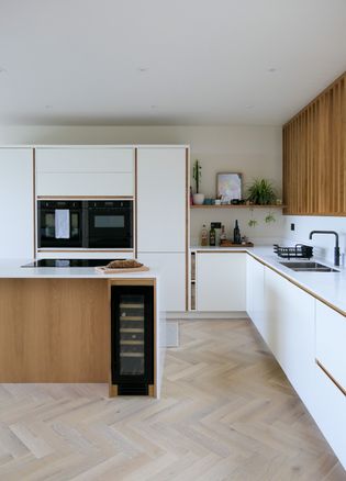 A modern kitchen featuring a large island with a wine cooler, white cabinetry, a built-in oven, and a wooden accent wall. The floor has herringbone wood patterns, and there are plants and decorative items on the shelves.