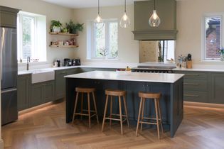 Shaker kitchen with green cabinetry, wood floors, and a blue central island with three stools. Pendant lights hang above. Bright and inviting atmosphere.