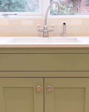 A shaker kitchen sink with a tall, polished silver tap is set against cream tile under a window. The light green cabinets add a calming touch.