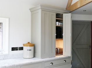 A kitchen with a light gray cabinet door open, revealing shelves with cups and a toaster. A crock labeled "EGGS" sits on a marble counter. Rustic, cozy feel.