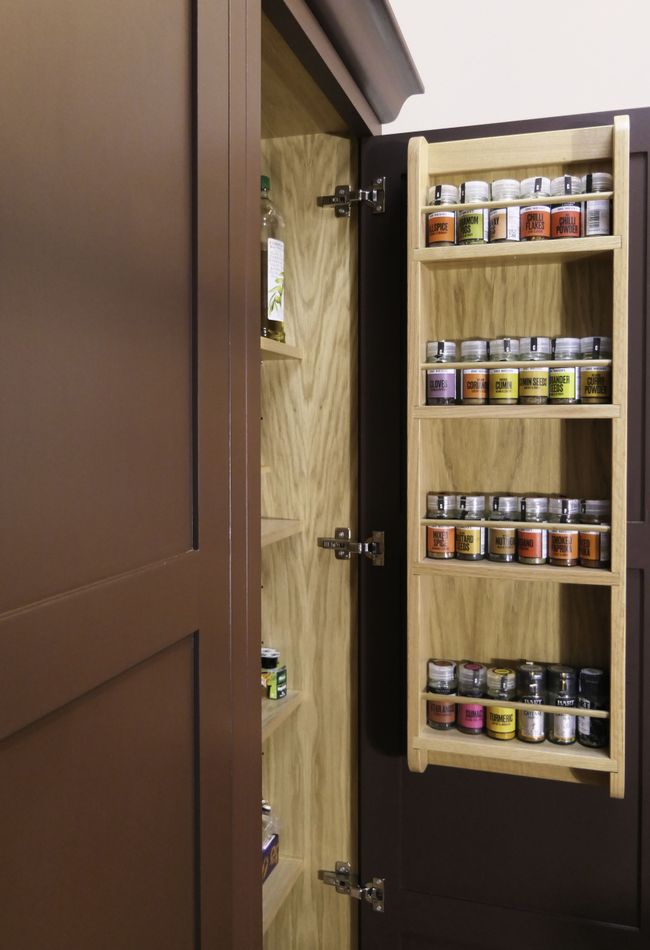 An open Shaker door to a larder with a handmade spice rack attached to it. Various different spices are on the rack with coloured labels.