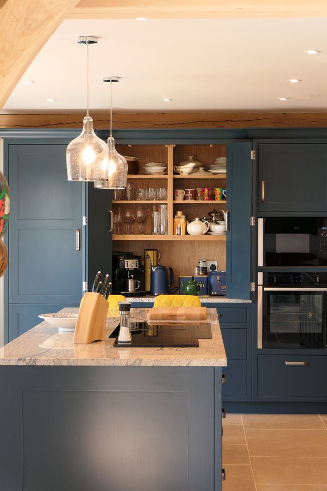 Spacious kitchen with blue cabinets, open pantry, and granite island. Hanging glass pendant lights add warmth. Modern and organized ambiance.