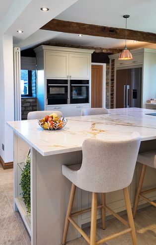 Shaker kitchen with a quartz island, upholstered bar stools, built-in ovens, a fridge, pendant light, and a bowl of fruit on the counter.