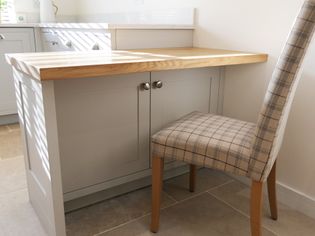 Shaker kitchen nook with a light wooden countertop, grey cabinets, and a plaid upholstered chair. Sunlight casts soft shadows.