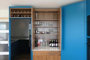 Open blue kitchen cabinet reveals a home bar with shelves of glasses and bottles. The modern design conveys a sleek, organized, and casual tone.