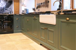 Modern kitchen with green cabinets, brass handles, and a white farmhouse sink. Black stove, marble countertops, and knives on a magnet strip.