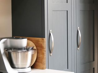 A shaker kitchen scene with a silver stand mixer next to a wooden cutting board on a white countertop. Gray cabinets with metal handles are in the background.