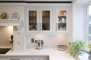 Bright kitchen with white cabinets, glass doors revealing dishes and glasses. A kettle and bowls sit on the counter. A plant adds a touch of nature.