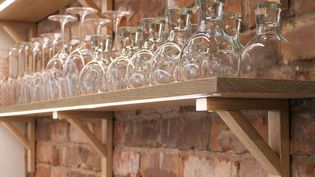 Wooden shelf against a brick wall holding rows of upside-down wine glasses. The setting is warm and rustic, evoking a cozy and inviting atmosphere.