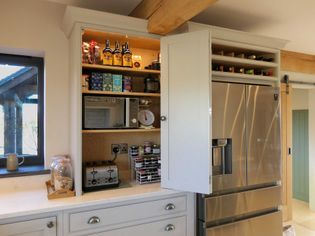 A shaker kitchen features an open cupboard with beverages, jars, a microwave, and toaster next to a large stainless steel fridge with a wine rack above.