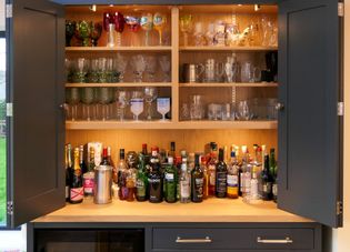 Open wooden liquor cabinet filled with assorted glassware and bottles with drawers and a fridge underneath.