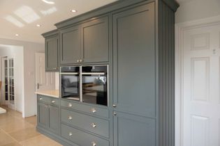 Elegant kitchen interior with tall, grey shaker-style cabinets. Built-in double oven, silver handles, beige tiled floor, and recessed lighting. Sophisticated ambiance.
