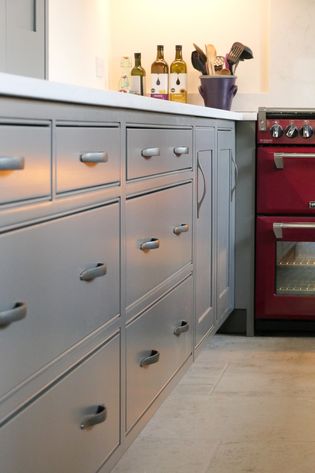 Shaker kitchen with grey cabinets, a red range oven, and utensils in a purple container on the counter with wine bottles are lined up near the backsplash.