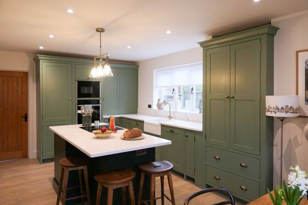 Green Shaker kitchen with lighter perimeter cabinetry and a darker island surrounded with wooden bar stools that has a fruit bowl and loaf of bread on top. White worktops throughout with built in double oven and farmhouse style sink.