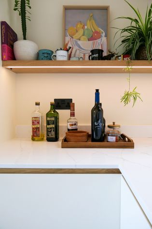 A modern kitchen countertop featuring various bottles of oil and beverages, a wooden tray with a bowl, and green plants hanging above.