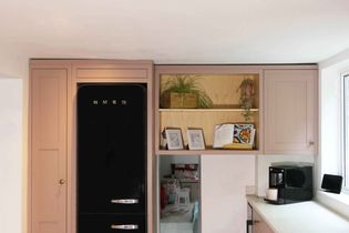 Shaker kitchen with a black SMEG fridge surrounded by pink cabinets. Open shelving displays plants and cookbooks. A coffee maker sits on the countertop.