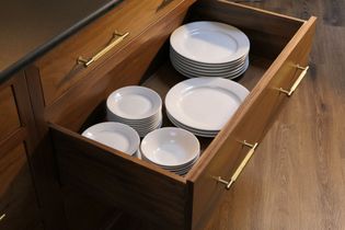 Open wooden drawer with neatly stacked white dishes, including plates and bowls, in a warm kitchen setting. The tone is orderly and inviting.