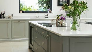 A shaker kitchen with light grey cabinets and a large island featuring white countertops. A vase of flowers provides a fresh touch. Natural light streams through a window, creating a bright, inviting atmosphere.