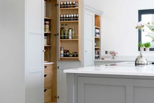 A shaker kitchen features grey cabinets with brass knobs. A pull-out shelf reveals mixing bowls, appliances, and utensils. The stove has a towel draped over the handle.