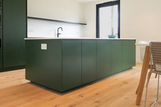 Modern kitchen with sleek dark green cabinetry and light wooden flooring. A black tap is on the white countertop. A bowl sits near a window, creating a fresh and minimalist atmosphere.