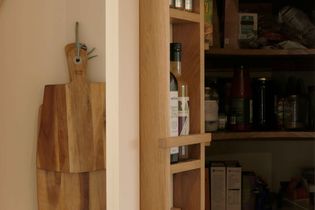 Wooden kitchen shelves with various items, including oil bottles and canned goods. A pair of wooden cutting boards hangs on the left wall.