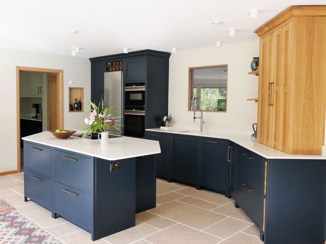 Spacious shaker kitchen with navy cabinets, wooden countertop cupboard and white worktops. A central island features a vase with flowers. A bright, clean atmosphere.