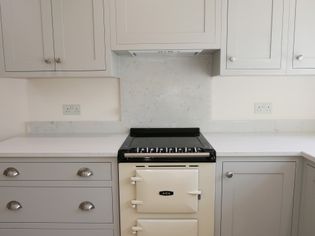 Shaker kitchen featuring a cream-colored AGA stove, surrounded by light grey cabinets and white countertops. The scene is minimalist and tidy.