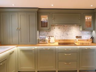 Shaker kitchen with muted green cabinets, wooden countertop, hexagonal tile backsplash, and under-cabinet lighting. A kettle and cutting board are on display.