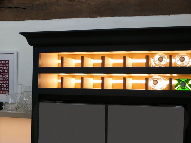 Illuminated shelving unit with warm lighting, featuring a modern black and wood design. Glassware and a framed poster are visible, evoking a cozy ambiance.