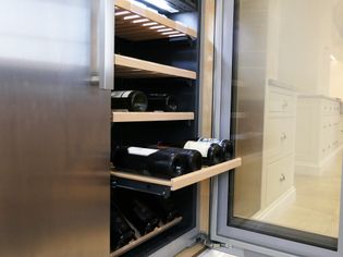 A sleek, open wine fridge with wooden shelves displaying bottles. The shaker kitchen setting exudes a clean, sophisticated ambiance.