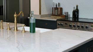 Shaker kitchen with a light worktop, brass taps, and a green glass bottle. Stainless steel stove and cutting board set in the background. Elegant and sleek.