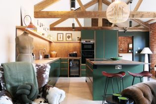 A stylish kitchen and living area with wooden beams, featuring a green kitchen, a cozy brown armchair, a dining table with white chairs, and decorative elements.