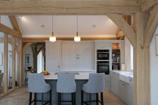 Shaker kitchen with exposed wooden beams, three gray barstools facing an island, pendant lights, and built-in ovens. Bright and inviting atmosphere.