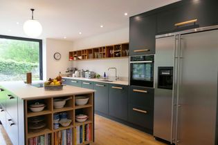 Modern kitchen with dark blue cabinets, stainless steel fridge, and a central island featuring a fruit bowl. Bright, with natural light from a large window.
