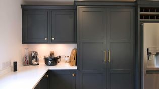 Shaker kitchen with dark cabinets and gold handles, featuring a white countertop. Appliances include a slow cooker and toaster; a stainless steel fridge is on the right.