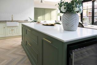 Elegant shaker kitchen with a quartz worktop island, green cabinets, gold handles, and a large vase with white flowers. Natural light adds a serene touch.