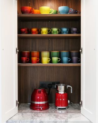 Open cupboard with neatly arranged rainbow-colored le cruset mugs on wooden shelves. Below, a red kettle and milk frother sit on a marble countertop.
