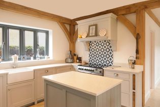 Rustic shaker kitchen with oak beams, grey cabinets, a central island, and geometric blue tiles. Natural light streams through large windows, creating a warm, inviting atmosphere.