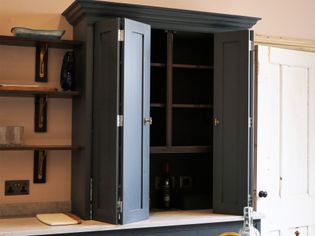 Open dark cabinet with shelves on a light wall, wooden shelves to the left holding a blue vase and glass items. A wine bottle is visible inside. Calm, organized.