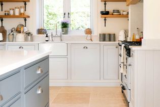 Bright kitchen with white cabinets, a farmhouse sink below a window, wooden shelves with jars and dishes, and a stove with a towel hanging. 