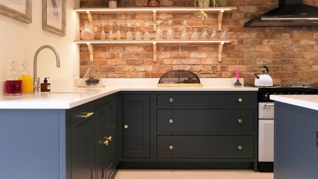 A shaker kitchen with dark grey cabinets, a brick backsplash, and white countertops. Shelves with glassware add elegance, creating a warm, inviting ambiance.