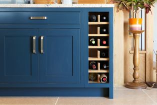 Blue kitchen cabinet with a built-in wooden wine rack holding nine bottles. Granite counter above, plant nearby, and warm, cozy tones.