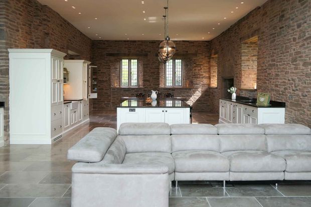 Open-plan Shaker kitchen and living room with stone walls, white cabinetry, a central island, large sectional sofa, windows, and a stone fireplace.