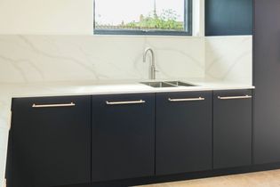 Modern kitchen with sleek navy blue cabinets and handles, white marble effect backsplash, and a stainless steel tap under a bright window.
