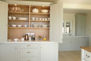 Open kitchen cabinet with neatly arranged white dishes, cups, bowls, and glassware on wooden shelves. A coffee maker and orange juice sit below. Calm, organized ambiance.