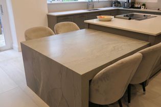Shaker kitchen with a large island, surrounded by beige velvet chairs. Light-coloured tiles and grey cabinets create a clean, spacious look.