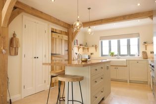 Bright kitchen with wooden beams, an island with two stools, pendant lights, cream cabinets, and large window. A rustic and inviting ambiance.